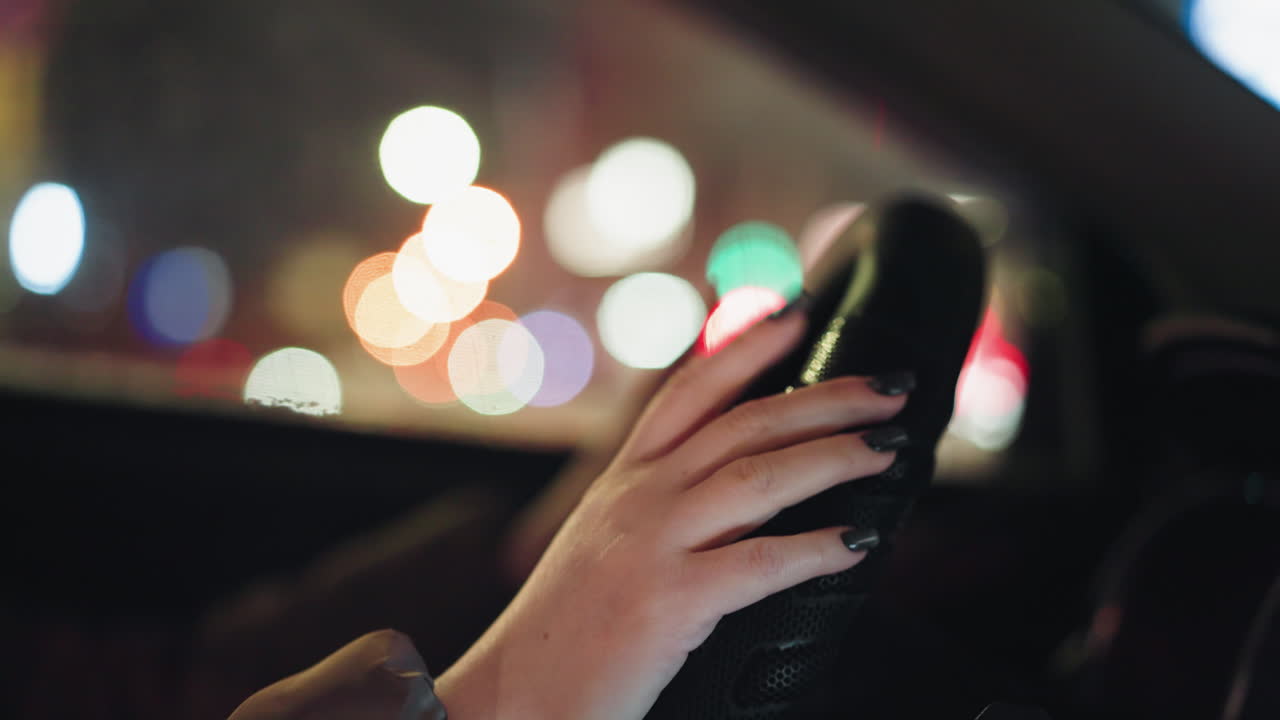 vista en primera persona de la mano de una mujer con uñas pintadas de negro sosteniendo un volante, capturada por la noche con luces borrosas de la ciudad en el fondo