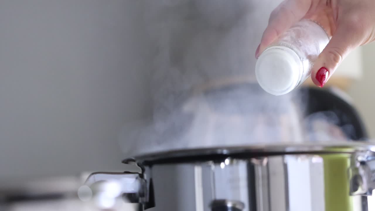A hand seasoning a pot with a shaker while steam rises