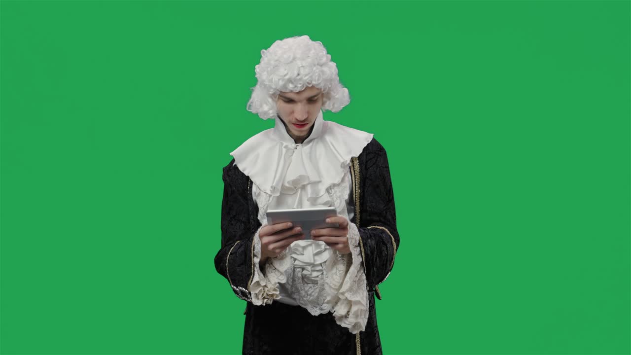 검은색 빈티지 슈트와 색 가발을 입은 궁정 신사의 초상화, 태블릿에서 게임을 하고 승리하고, 녹색 화면 배경으로 스튜디오에서 포즈를 취하는 젊은이.