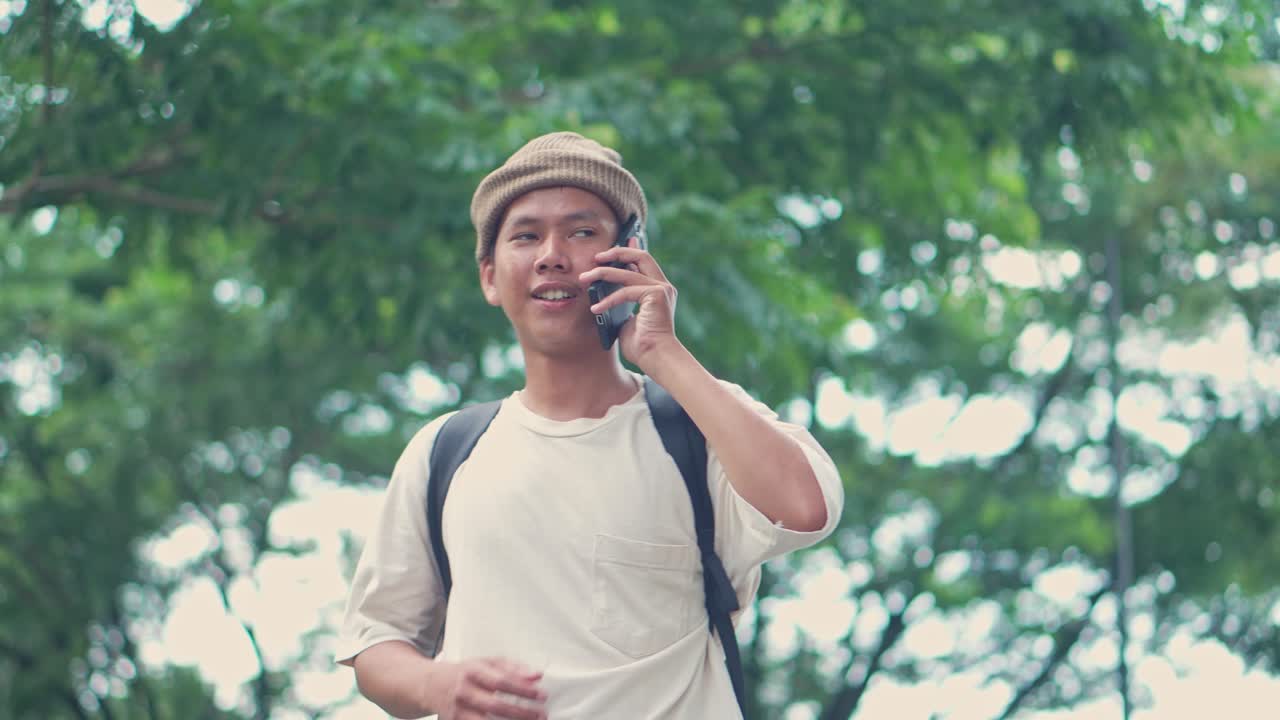 Young Asian College Student Talk On The Phone And and Raises His Hand To Indicate His Location To His Friends In The Park.