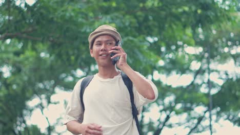 young asian college student talk on the phone and and raises his hand to indicate his location to his friends in the park.