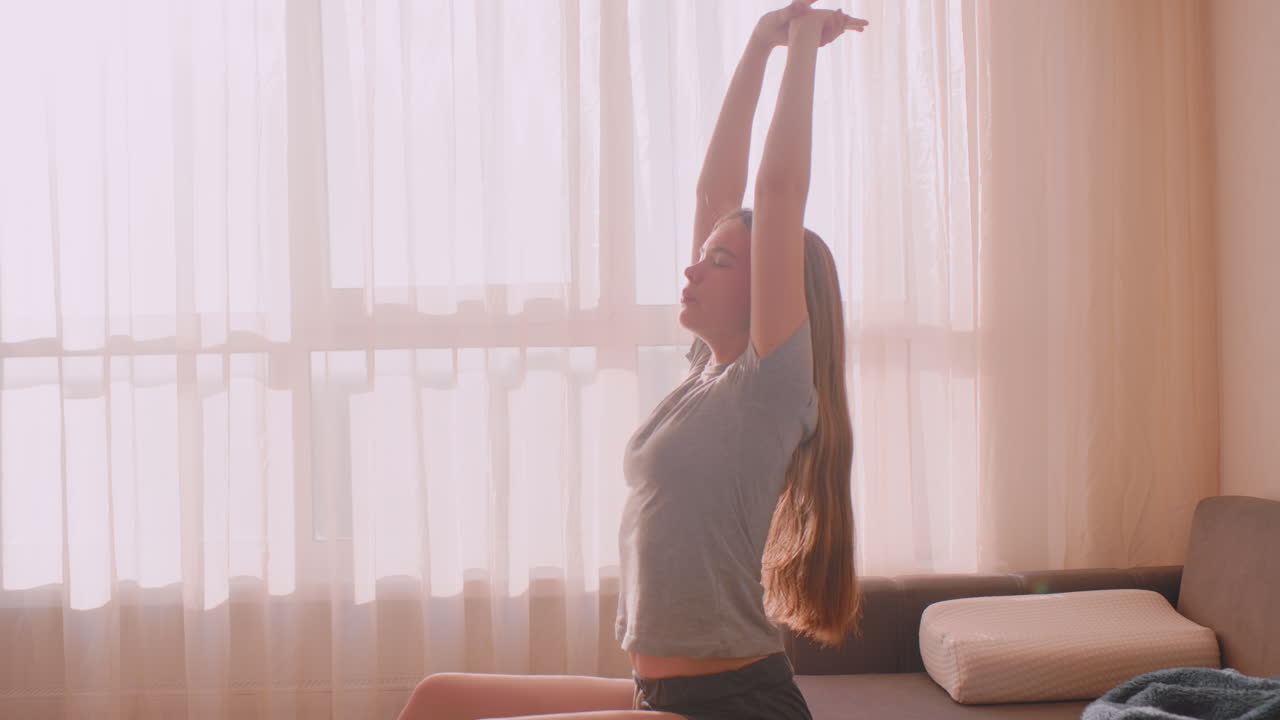 Young remote designer sits near curtain-covered window gently stretching shoulders to release tension after long working hours, bathed in soft golden morning sunlight in cozy home setting