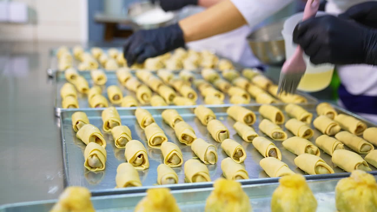 Three big baking sheets full of croissants are on the metal table. Baker's hand cover the desserts with egg. Close up.