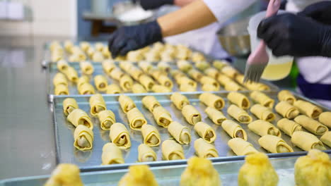 three big baking sheets full of croissants are on the metal table. baker's hand cover the desserts with egg. close up.