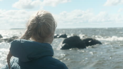 primo piano di una ragazza bionda in piedi sulla spiaggia vicino al mare, guardando le onde, due kayak sdraiati sulla spiaggia, una ventosa e fredda giornata estiva in finlandia, vicino a vaasa