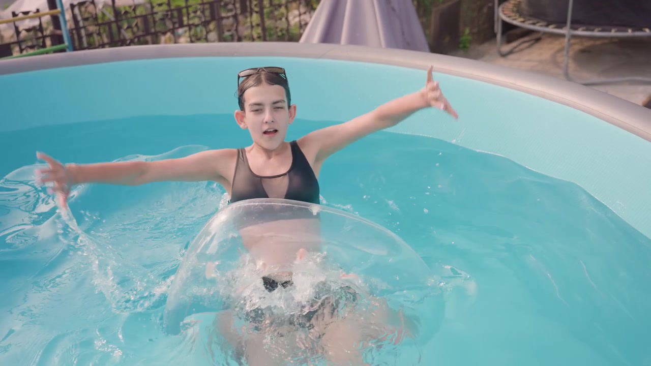 Young girl enjoys time in small above ground pool wearing swimwear and inflatable ring. Girl moves through water sometimes holding onto or playing with floatation device while surrounded