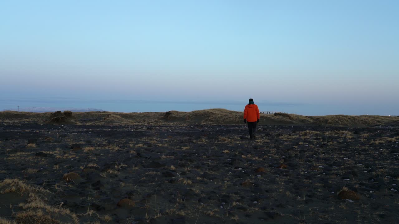 주황색 겨울 재킷을 입은 남자가 바다 해안선을 향해 걸어갑니다 - 정적 샷