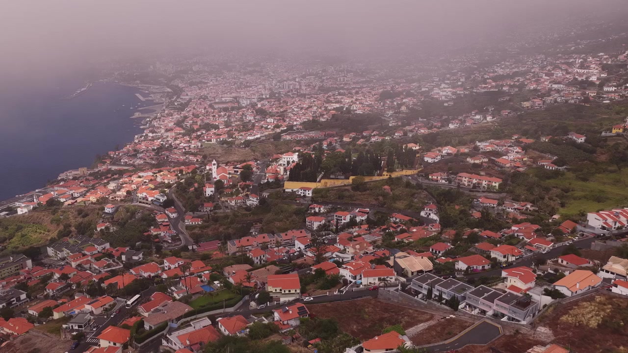 Funchal'ın Terakota Çatılarını ve Yumuşak Bir Pusla Örtülü Kıyı Şeridini Yakalayan Kapsamlı Drone Videosu. Geniş Hava Çekimi, Kentsel Peyzajı, Atlantik Kıyısını ve Kasvetli Bulutlu Aydınlatmayı Vurguluyor
