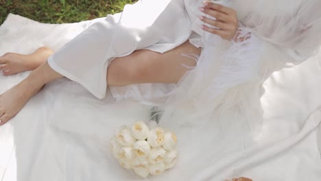 bride sitting on the grass in a white dress with champagne and bouquet