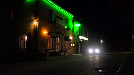 Ein-Auto-Fährt-Nachts-An-Einem-Kleinen-Französischen-Hotel-Mit-Blinkendem-Neonschild-Vorbei