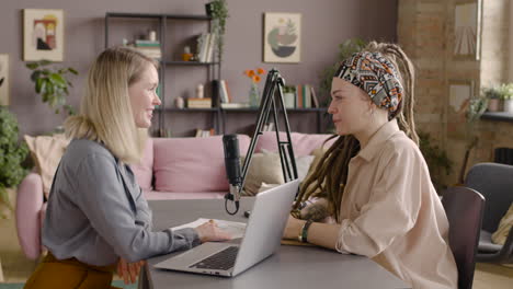 side view of two women recording a podcast talking into a microphone sitting at desk with laptop and documents