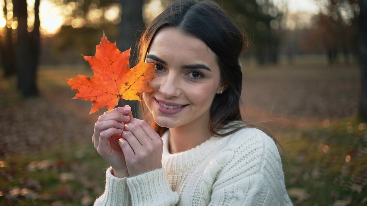 Fotografo che chiama una donna con un maglione bianco, che tiene una foglia d'acero arancione verso l'obiettivo, in posa in un parco.