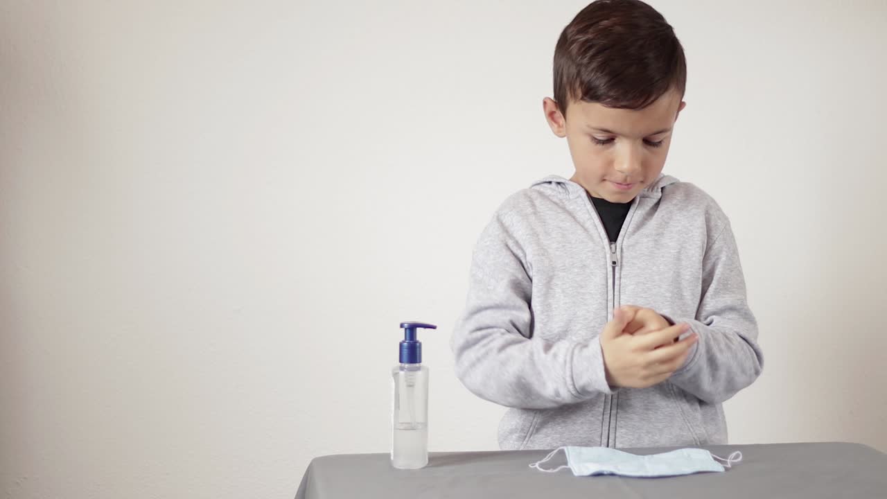 회색 재킷을 입은 소년은 손에 알코올을 바르고 안면 마스크를 착용함으로써 적절한 위생을 알고 있습니다.