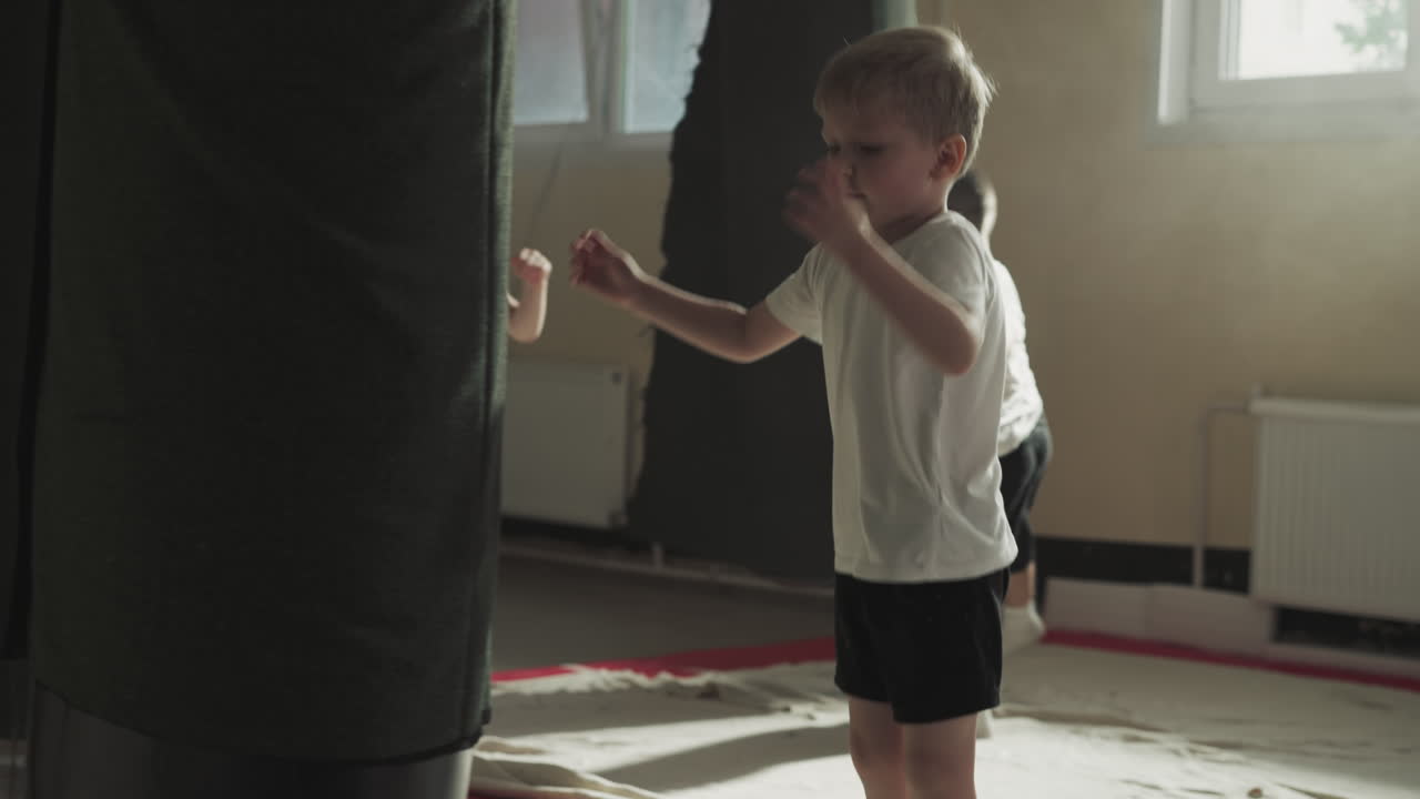 Focused child kicks huge punching bag in sunny gym. Little boy trains beating hanging equipment with foot in martial arts school slow motion. Cute little fighter