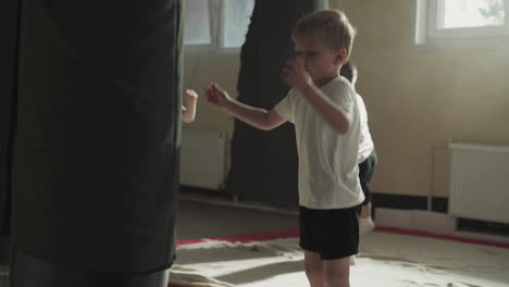 focused child kicks huge punching bag in sunny gym. little boy trains beating hanging equipment with foot in martial arts school slow motion. cute little fighter