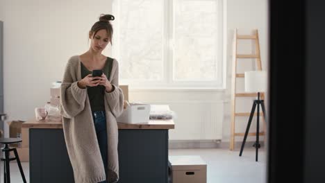 joven mujer caucásica usando un teléfono móvil mientras desempaqueta cosas en una casa nueva