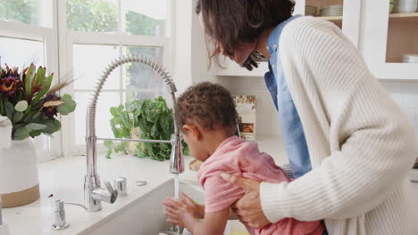 madre milenaria de pie en el fregadero de la cocina ayudando a su hijo a lavarse las manos, vista lateral, de cerca