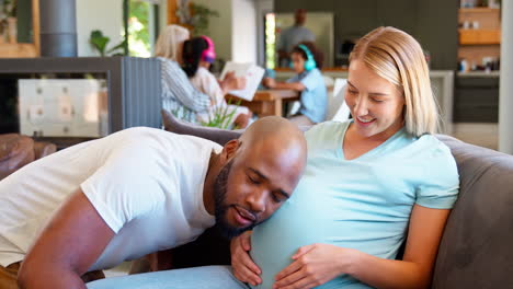 couple with man listening to pregnant woman's stomach on sofa at home with multi-generation family