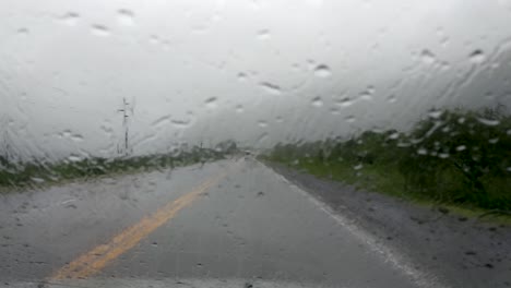 Raindrops-on-the-windshield-while-driving-in-heavy-rain,-low-grey-clouds-hang-above-the-wet-road