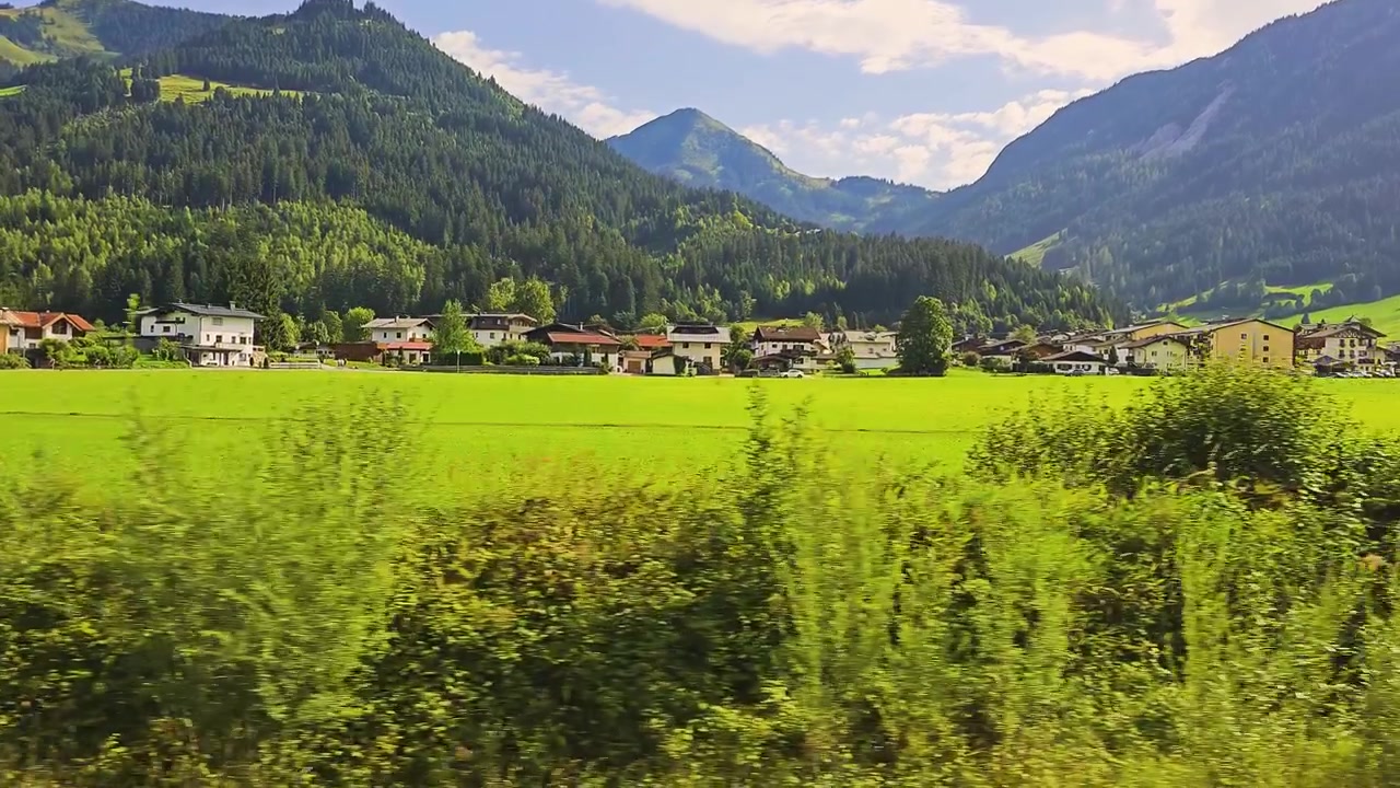 Paesaggio estivo da un treno in movimento in Austria, con pascoli verdi, piccole fattorie, villaggi di montagna e colline coperte di pini sotto un cielo alpino di un blu brillante