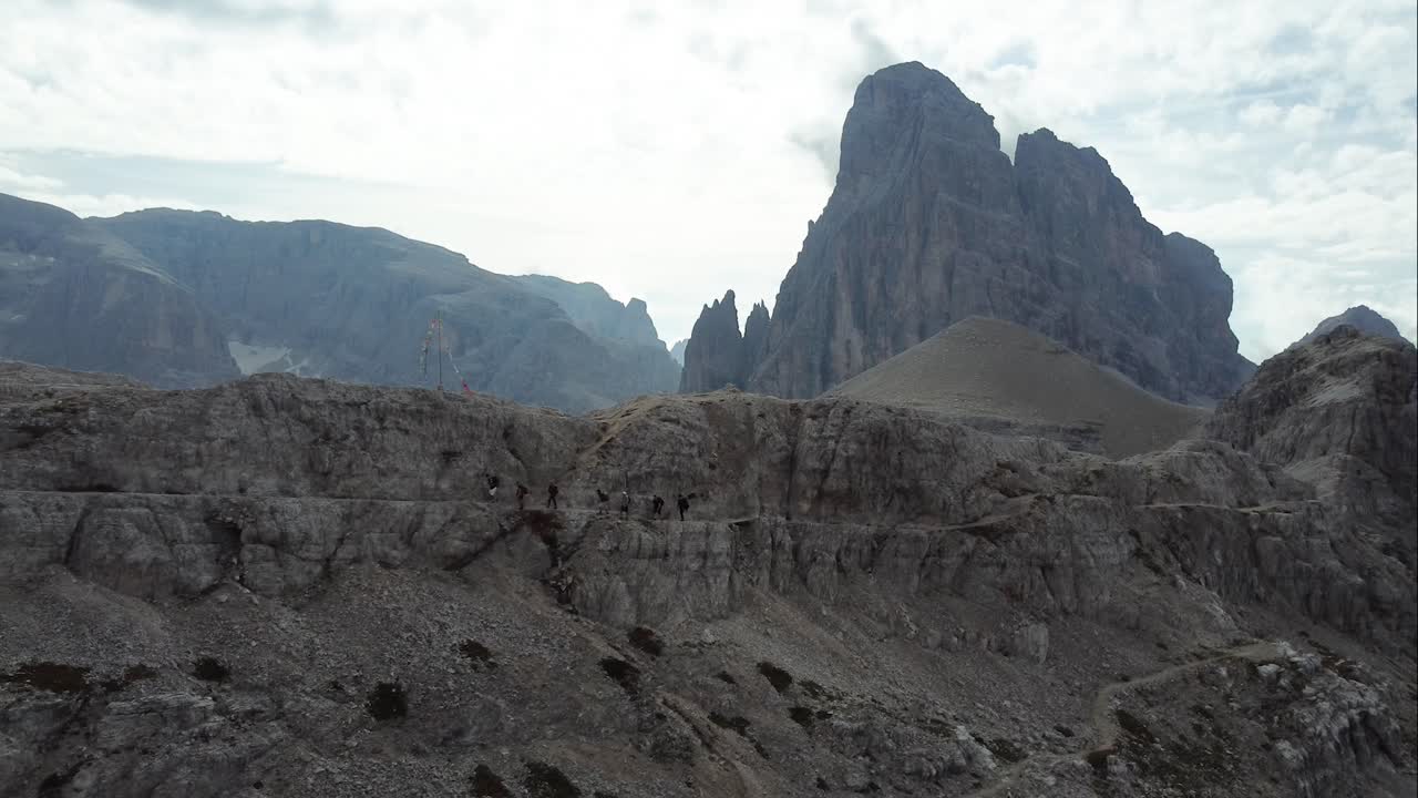 7 excursionistas caminan a un lado de un sendero de montaña en un acantilado empinado con formaciones rocosas sobresalientes y el increíble paisaje de los dolomitas en el fondo en el norte de italia