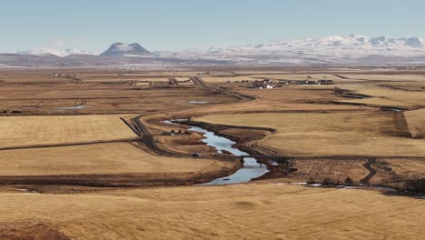 Drone flying over Þjórsá valley in Iceland, revealing winding rivers through golden fields and distant snow-covered mountains under a clear winter sky.