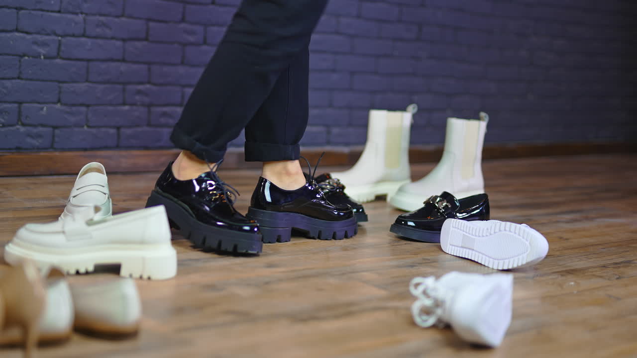 Trying on massive black shoes on tractor soles. Female feet pose among the diverse footwear scattered by the floor. Blue wall backdrop.