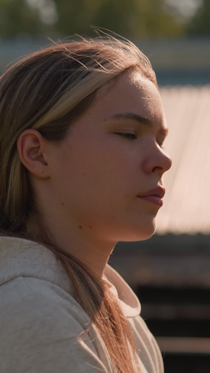 una mujer joven con una sudadera de color ceniza con el cabello atado hacia atrás camina afuera mientras la luz del sol se refleja en su cara, proyectando un brillo cálido, el fondo muestra los asientos vacíos del estadio borrosos