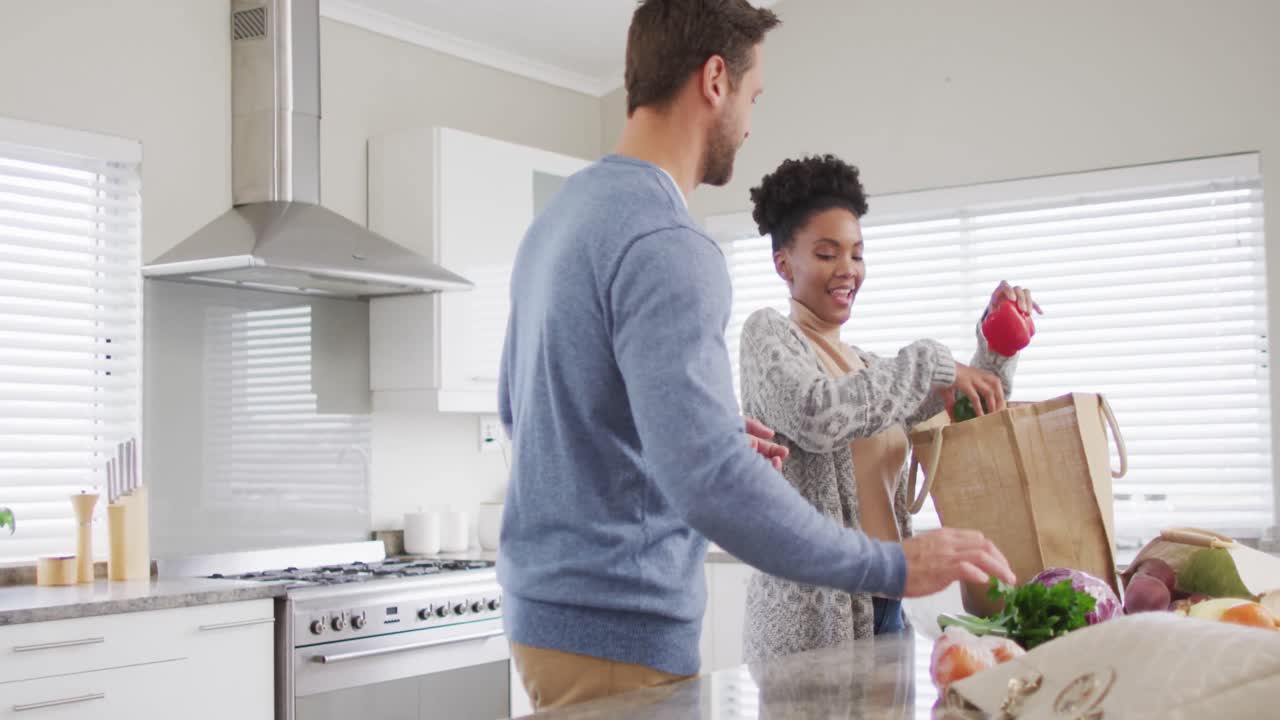 Video of happy diverse couple unpacking groceries in kitchen