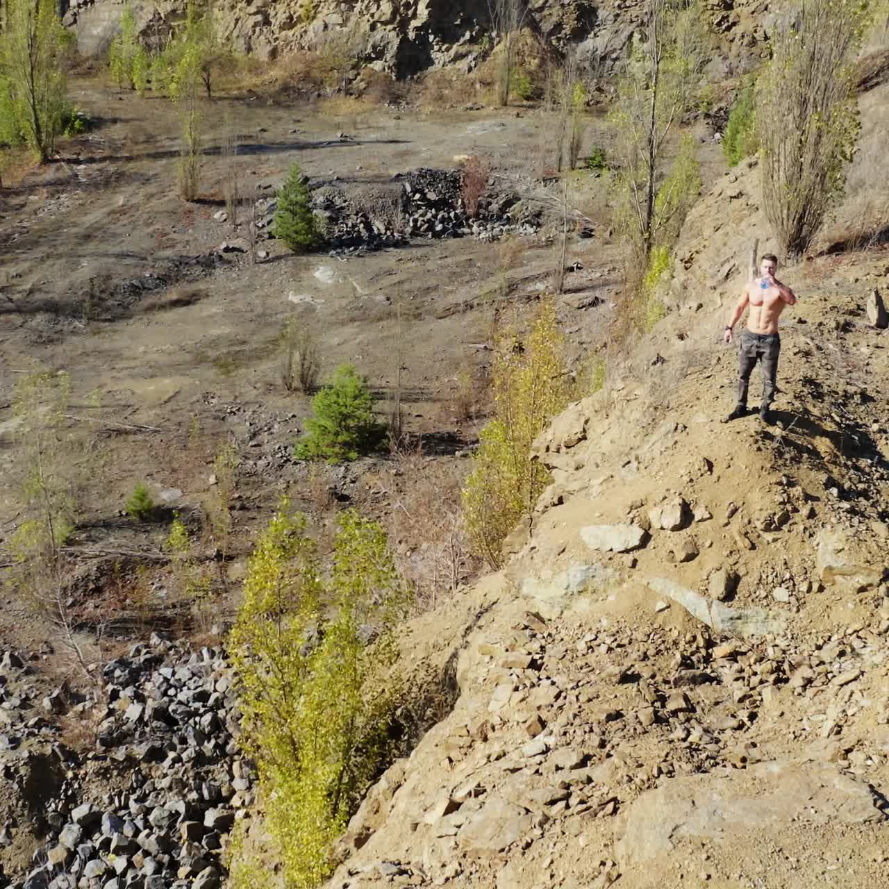 Young sportsman with a bottle of water in nature. Shirtless athlete drinking water after the workout in the mountainous area. Aerial view.