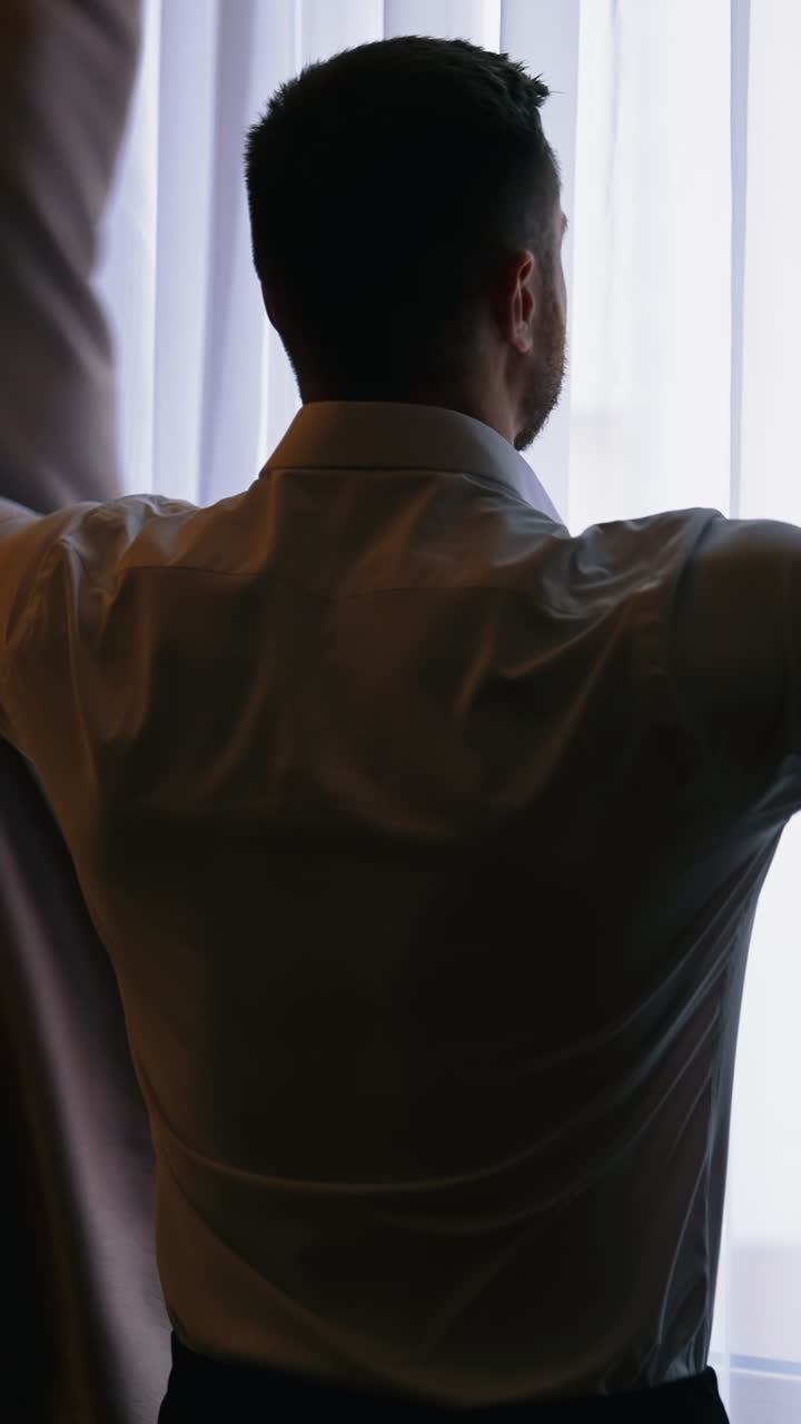 Young man opening curtains at hotel room. Man in white shirt opening the dark window curtains in a room. Relaxed man looking on the window. Back view. Vertical video