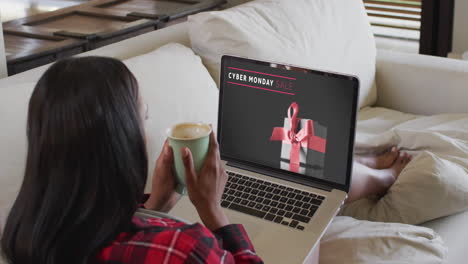 biracial woman on couch with coffee using laptop for online shopping, slow motion