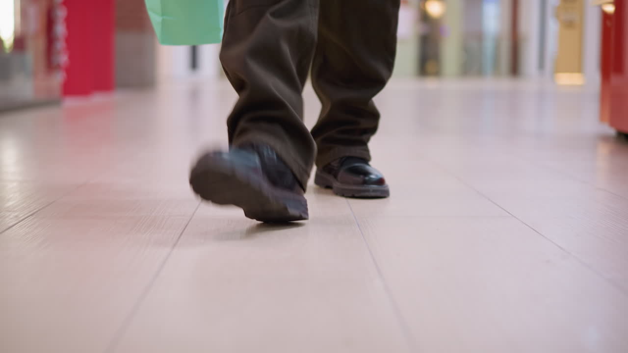 low angle of legs and shoes of person walking on polished tile floor in shopping mall carrying green bag visible top left blurred store lighting reflections motion clip casual pant denim fashion style