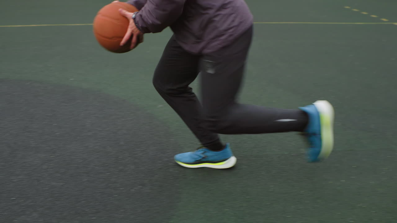 Genç bir oyuncu açık hava sahasında basketbol topu sektiriyor; arka planda şehir silüeti ve apartman blokları, dokulu asfaltı çizen spor ayakkabılar, top potaya doğru sekip yay çiziyor, çemberden gelen bir ses