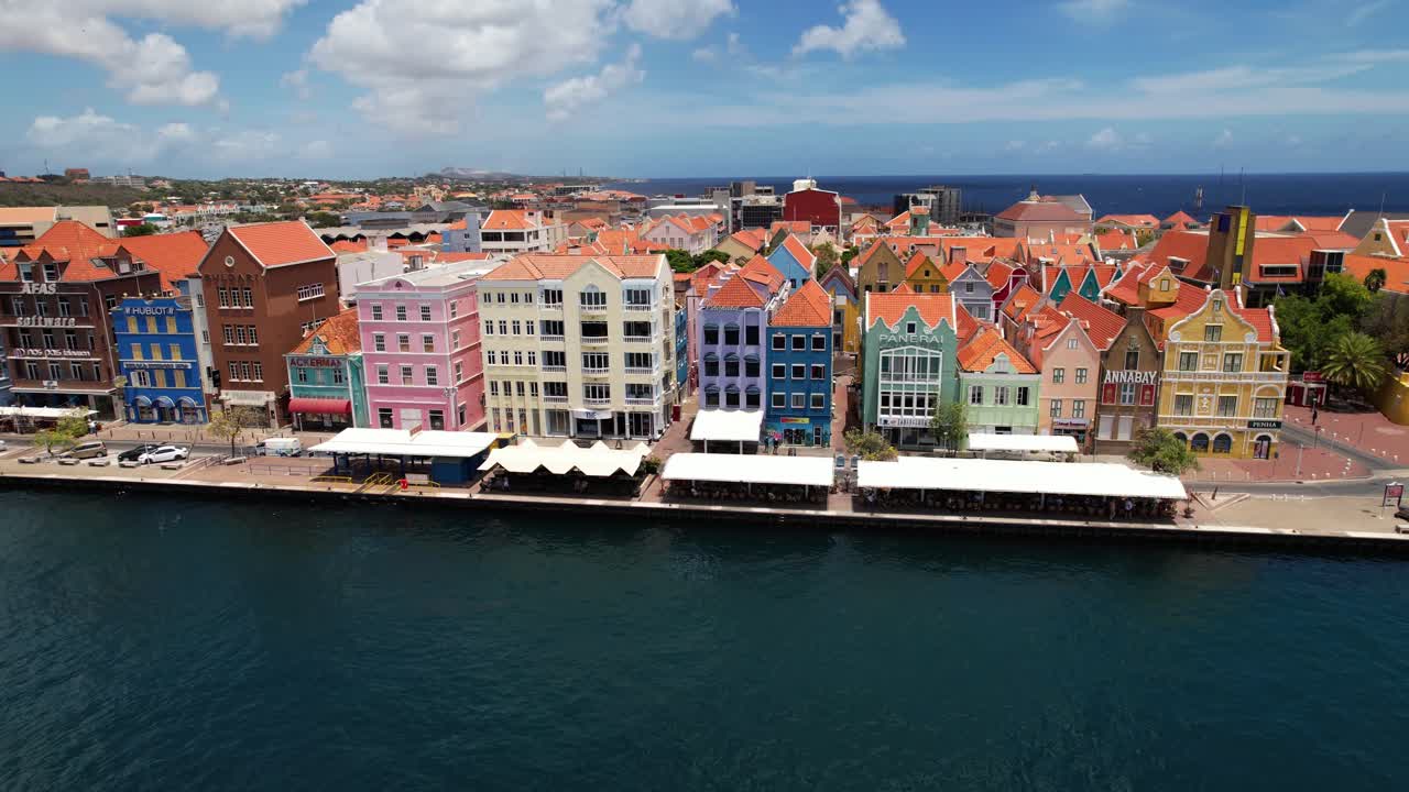 Aerial drone footage showcases Willemstad, Curacao, in a magnificent circle pan. Colorful buildings line the promenade on a sunny summer day, capturing slow city life with few cars and pedestrians.