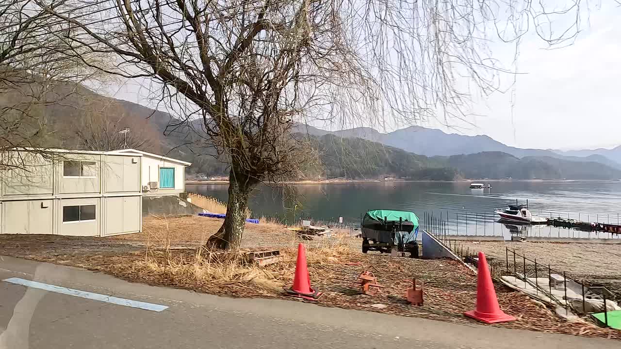 A serene drive by Lake Kawaguchi, capturing tranquil waters, distant mountains, and sparse roadside buildings under soft daylight