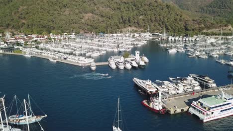 turkey, marmaris, drone footage on a sunny day over the marina, mega yachts, sailboats moored, a sailboat is approaching to the entrance of the marina, camera turns towards to the river and the town.