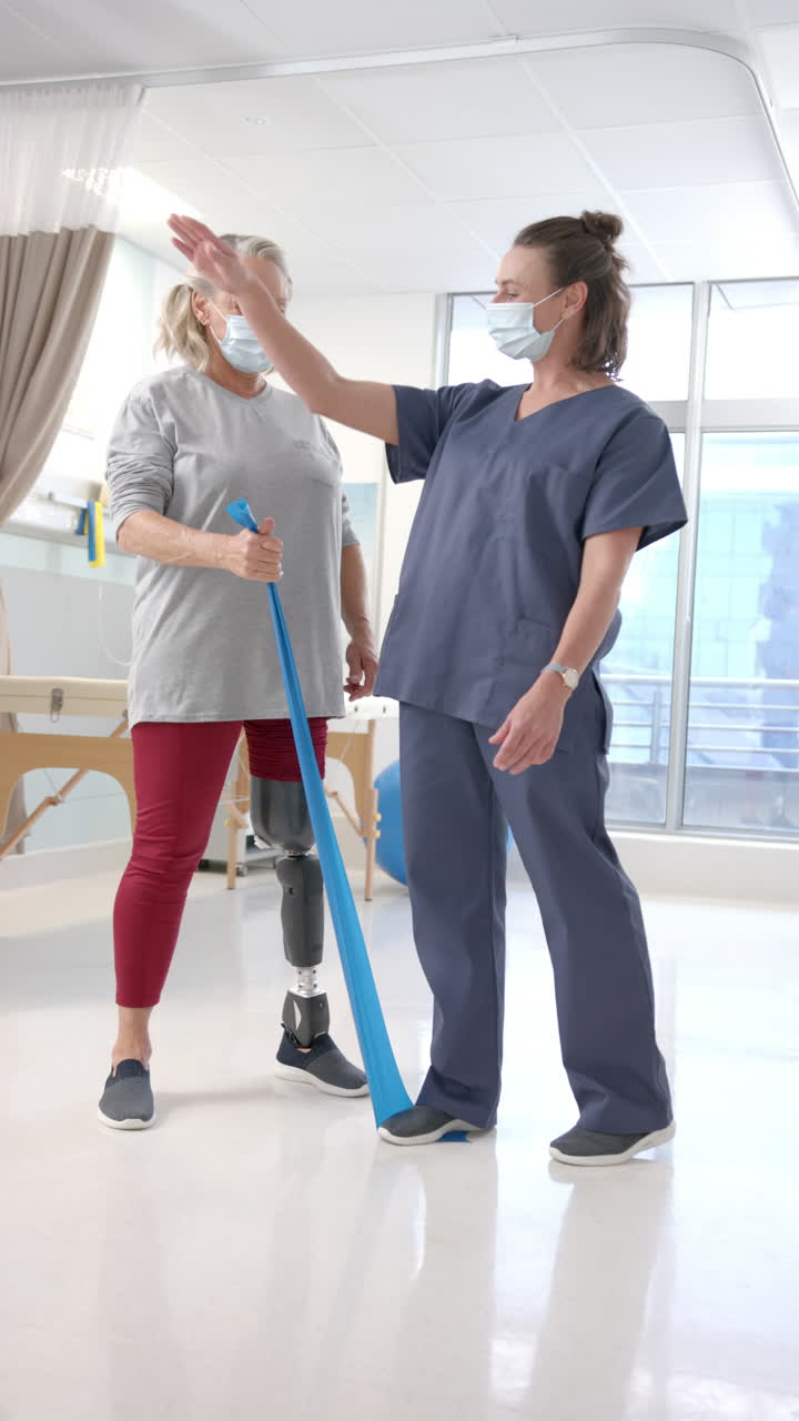 Vertical video of caucasian female physiotherapist helping female patient to exercise with bands