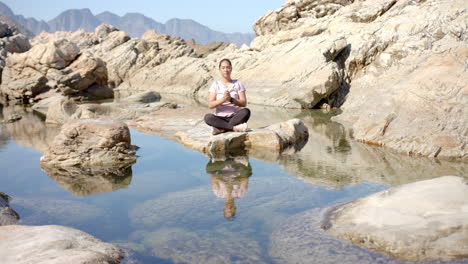 meditating on rock by mountain lake, woman enjoying peaceful outdoor setting