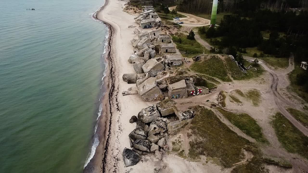 vista aérea del edificio de fortificación costero abandonado en los fuertes del norte de karosta en la playa del mar báltico en liepaja en un día nublado de primavera, turbina eólica, tiro amplio de drones moviéndose hacia atrás, inclinado hacia arriba