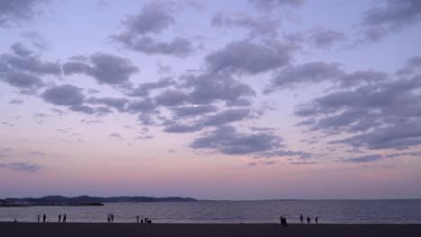 Amplia-Vista-Abierta-En-La-Hermosa-Playa-Al-Atardecer-Con-Nubes-Altas-Y-Luna-Y-Siluetas-De-Personas-Caminando-En-La-Arena