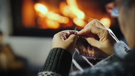 close-up of the hands of an elderly woman who knits a dull thing on the background of the fireplace