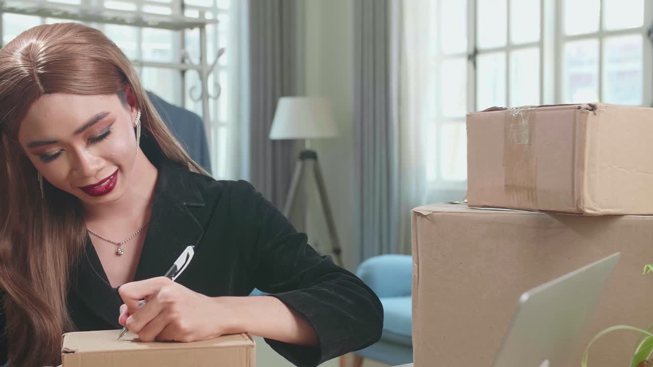Young Asian Transgender Woman Business Owner With Boxes On The Table Happy Online Sales Job, Get An Order From Customers, Take Notes On The Box, And Make Arrangements For Delivery By Post