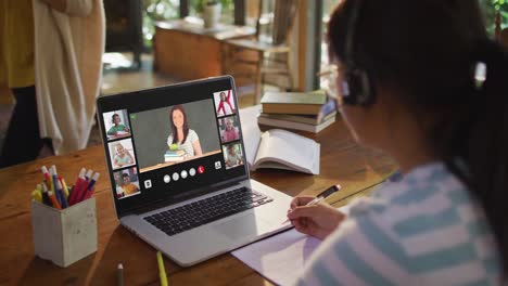 Schoolgirl-using-laptop-for-online-lesson-at-home,-with-diverse-teacher-and-class-on-screen