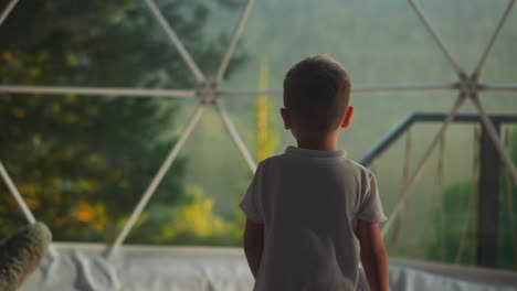 pensive kid examines landscape. preschooler boy approaches big sunlit window in comfortable tent slow motion backside view. calm child admires nature closeup on blurred background