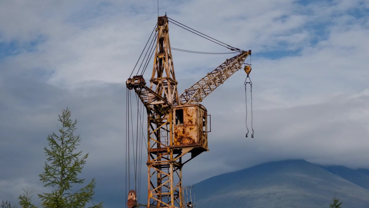 Una gru arrugginita e ferma si erge immobile contro montagne distanti e nuvole alla deriva, con una struttura metallica invecchiata e cavi pendenti incorniciata da alberi immobili.