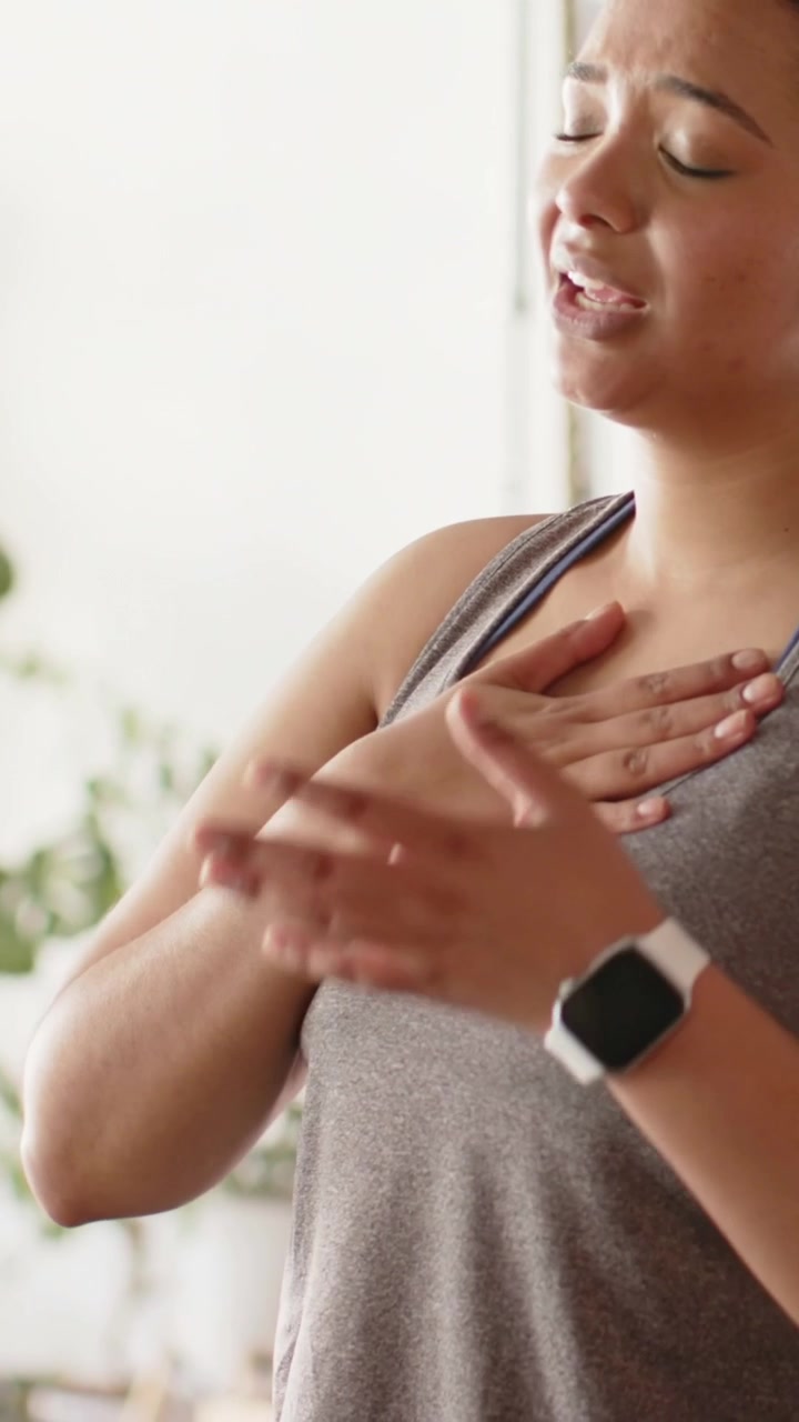 Vertical video: Raising right arm, woman in gray tank top addressing off-camera, touching chest