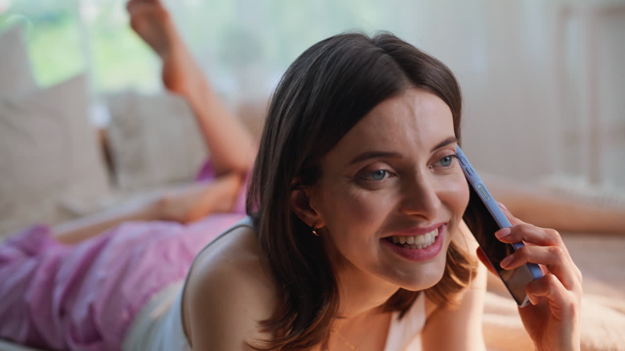 Close-up de uma garota atendendo uma chamada no celular enquanto relaxa na cama de um apartamento. Mulher sorrindo