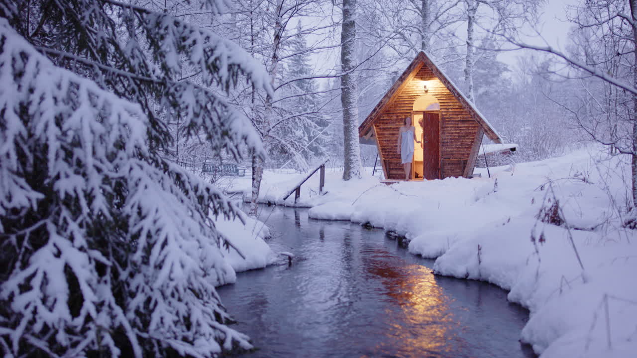 Couple sits outside sauna surrounded by snowy winter wonderland, view over river
