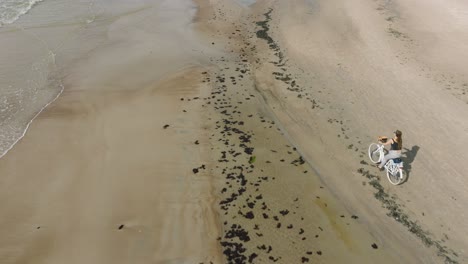 vista aérea con una joven de cabello largo montando una bicicleta en la playa de arena, día soleado, playa de arena blanca, concepto de estilo de vida activo, amplio tiro de drone de ojo de pájaro moviéndose hacia adelante